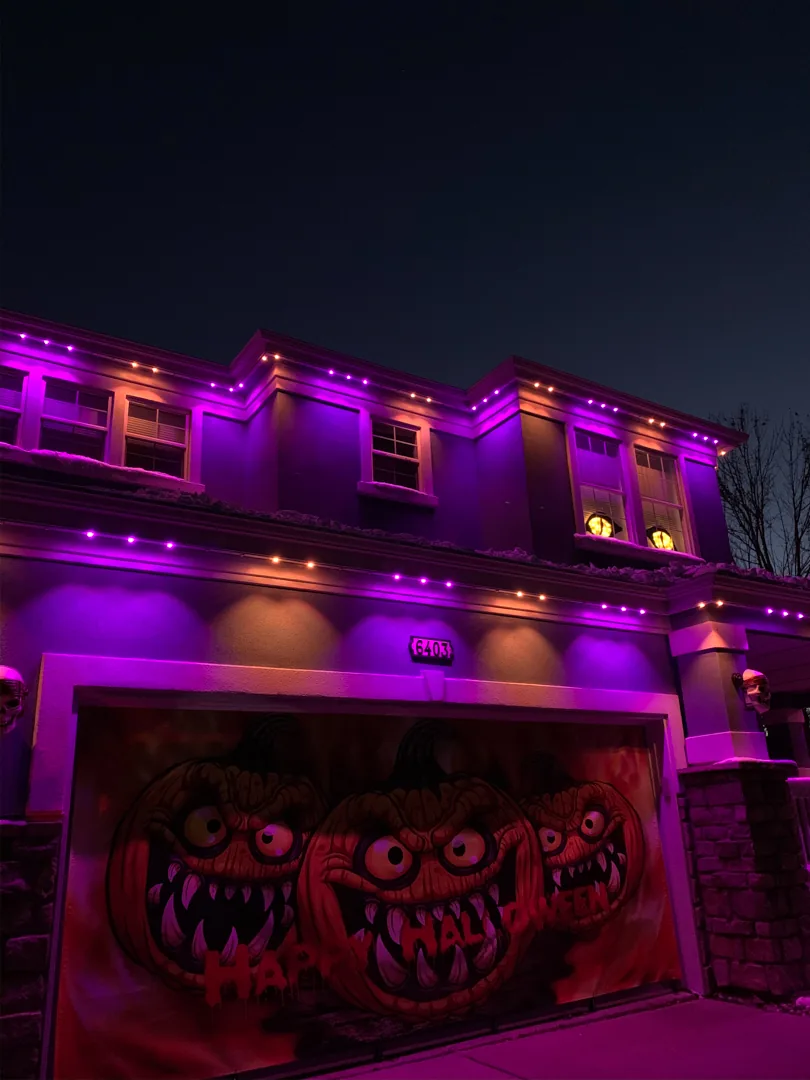 Two-story home with its roofline lit orange and purple for Halloween, monster mural on the garage