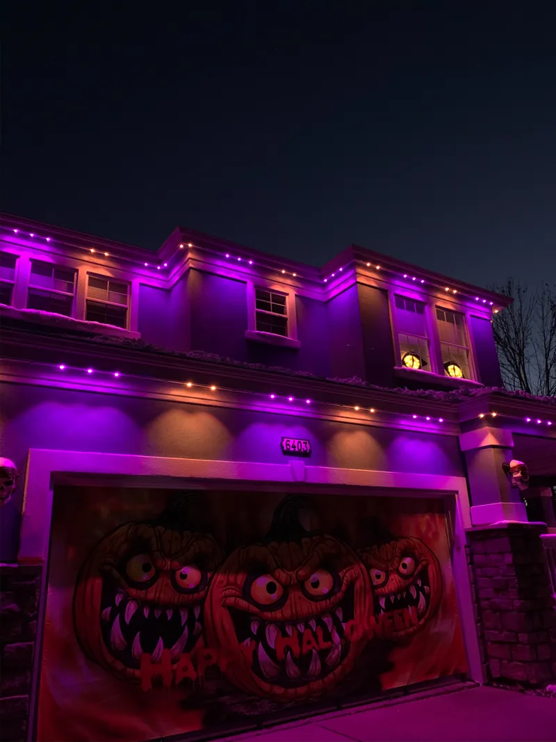 Two-story home with its roofline lit orange and purple for Halloween, monster mural on the garage